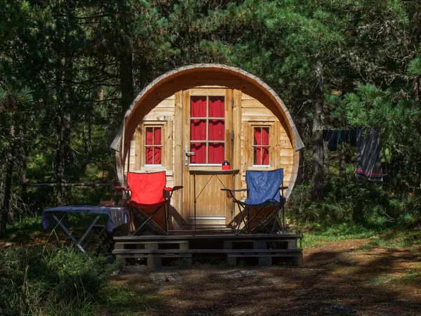 A round timber hut with red curtains and two camping chairs on its deck, among pine trees