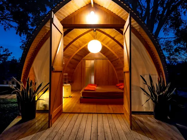 An arched wooden pod at dusk with its doors open, showing a lamp-lit bed inside