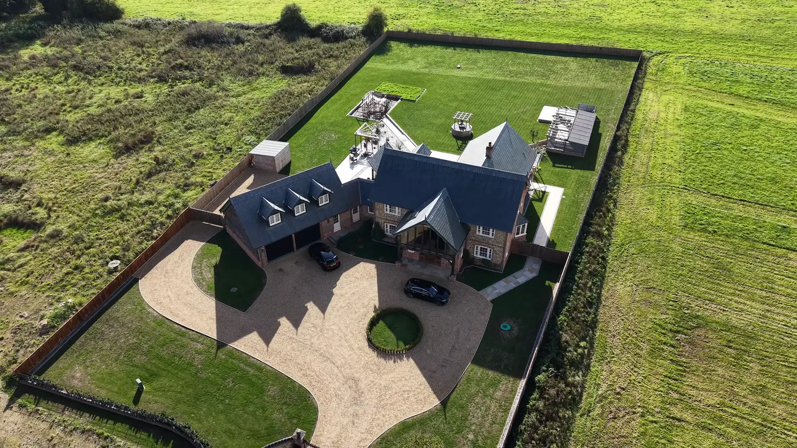 Aerial view of a new flint house with slate roofs, an oak porch, a double garage and a gravel drive, surrounded by fields