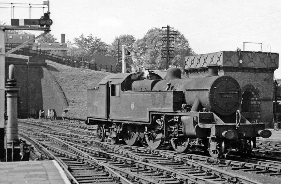 Black-and-white photo of a steam locomotive at Hibel Road Station, with a signal gantry and stone bridge behind